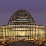 A highly detailed, photorealistic depiction of the European Parliament building in Brussels at dusk, with the iconic dome illuminated by soft blue LED strips, surrounded by a bustling crowd of diverse professionals in business attire holding digit…