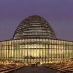 A highly detailed, photorealistic depiction of the European Parliament building in Brussels at dusk, with the iconic dome illuminated by soft blue LED strips, surrounded by a bustling crowd of diverse professionals in business attire holding digit…