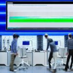 A modern server room with rows of humming racks, each rack illuminated by soft blue LEDs. In the foreground, a diverse team of developers in casual attire stands around a large transparent screen displaying a real-time CO₂ emission dashboard. The …