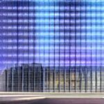 A photorealistic depiction of the German Bundestag building at dusk, illuminated by soft blue LED strips, with translucent holographic data streams flowing from the building into the surrounding cityscape, showing digital connections to cloud serv…
