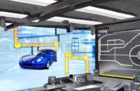 Techniker in blauem Overall in einem industriellen Kontrollraum, der an einem Hebel arbeitet, während ein großer Digitalbildschirm eine Echtzeit-Simulation des Bremssystemes eines Fahrzeugs mit ABS-Schema an der Wand zeigt.