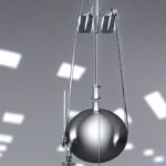 Ein klassisches Labor-Setup mit einem glänzenden Metallpendel, das an einer rostfreien Stahlstange hängt, steht auf einer weißen Arbeitsfläche. Im Hintergrund ein großes, transparentes Messgerät mit einer Skala von 0 bis 10 Newton, das die Kraftme…